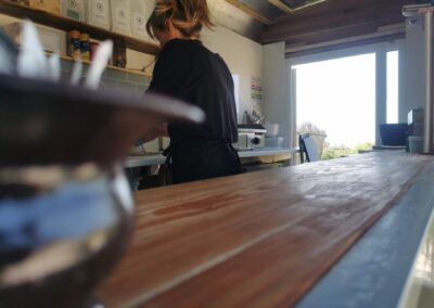 Barista working inside a rustic trailer café with a wooden counter, eco-friendly coffee supplies, and sunlight streaming through an open hatch.