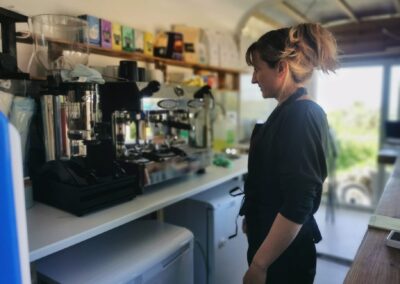 Barista standing beside an espresso machine in a bright, eco-friendly café trailer with sustainable products on the shelves.