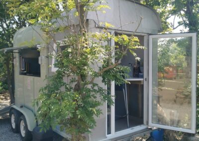 "Converted trailer café with open door and serving hatch, nestled under trees at a peaceful campsite with gravel and natural landscaping."