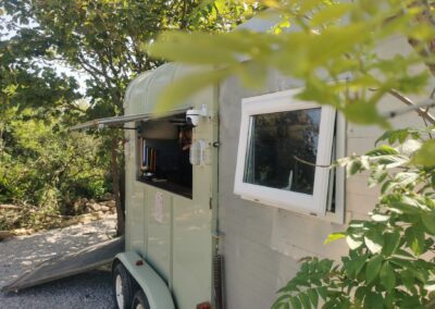 Eco-friendly trailer café with open serving hatch, parked among trees and greenery at a quiet sea-side campsite.