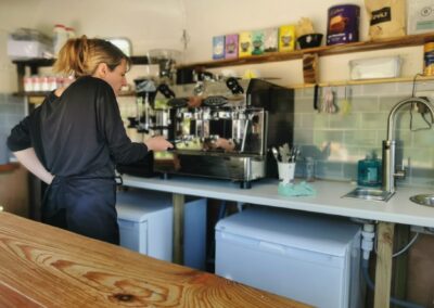 "Barista preparing coffee in an eco-friendly trailer café with a professional espresso machine and ethically sourced products on shelves."