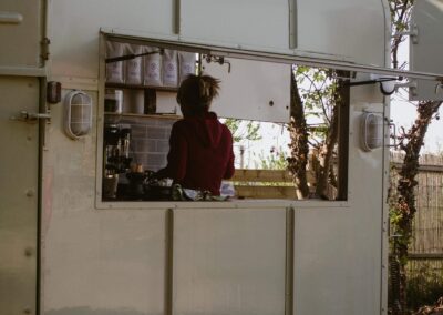 "Barista working inside a converted trailer café with open serving hatch, nestled among trees and greenery at a rural campsite."