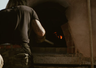 "Person tending the fire inside a rustic dome-shaped wood-fired oven, with flames glowing and firewood stacked below in an outdoor kitchen."