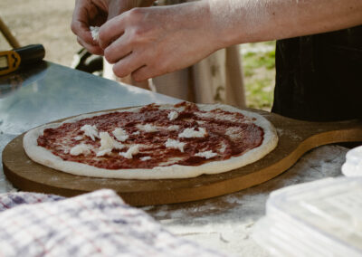 "Hands topping a pizza with fresh cheese in an outdoor kitchen setup, on a flour-dusted surface at a campsite."