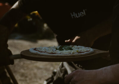 "Uncooked pizza with tomato, cheese, and basil on a wooden peel, held by a chef in a black apron near a wood-fired oven."