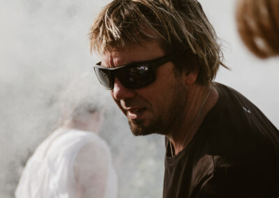 "Close-up of a man in sunglasses surrounded by smoke, focused while cooking outdoors near a pizza oven."