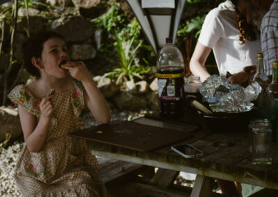 "Young girl in a floral dress eating pizza at an outdoor wooden picnic table after a shared meal."