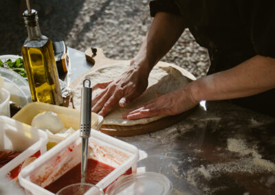 "Close-up of hands shaping pizza dough on a floured board, surrounded by tubs of tomato sauce, mozzarella, and fresh ingredients."
