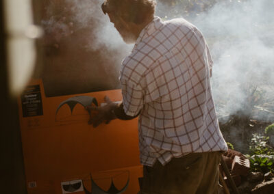 "Man tending to a smoking outdoor oven made from natural materials, wearing shorts and a checkered shirt in a rustic, sunlit setting."