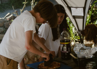 "Children gathered around a picnic table under a canopy, slicing a wood-fired pizza while a dog watches nearby."