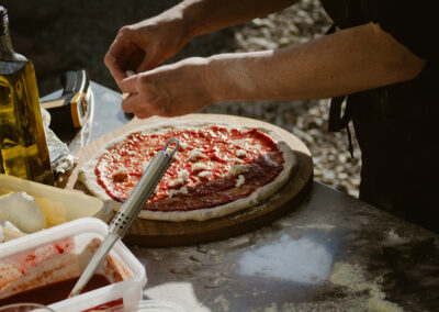 "Close-up of hands adding cheese to a tomato-covered pizza dough on a wooden board, surrounded by tubs of fresh ingredients and olive oil."