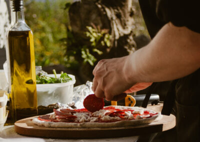 "Chef’s hand placing pepperoni slices on a pizza outdoors, with olive oil, rocket, and fresh toppings on a flour-covered table."