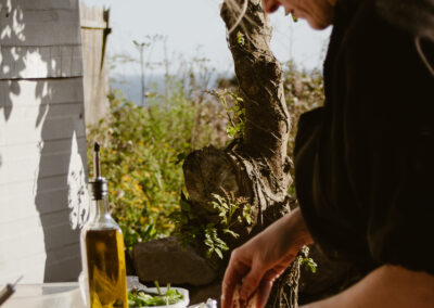 "Chef adding toppings to a pizza on a wooden peel outdoors, with fresh ingredients and olive oil beside a tree and sea view."