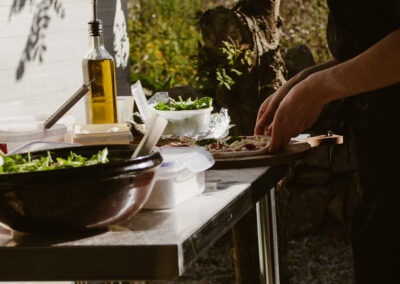 "Chef preparing pizza on a flour-dusted table with fresh toppings outdoors, beside a tree and natural foliage in golden sunlight."