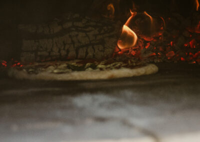 "Raw pizza baking beside glowing embers and flames inside a wood-fired clay oven."