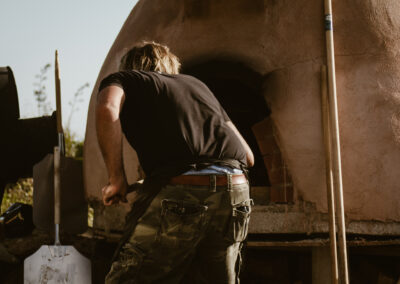 "A person in a black shirt and camouflage trousers leans into a clay pizza oven, using a peel to manage the fire inside during outdoor cooking."