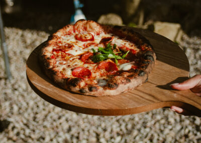 "Freshly baked pepperoni and mozzarella pizza topped with rocket leaves, held on a wooden board outdoors."
