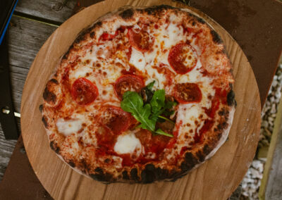 "Top-down view of a wood-fired pepperoni pizza with melted mozzarella and rocket leaves, served on a wooden board outdoors."