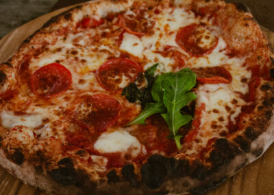 "Close-up of a rustic wood-fired pepperoni pizza with melted mozzarella, tomato sauce, and fresh rocket leaves on a wooden board."