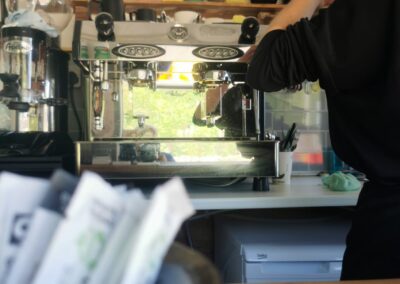 Barista working at an espresso machine in a sustainable trailer café, with Fairtrade sugar sachets in the foreground and eco products on shelves.